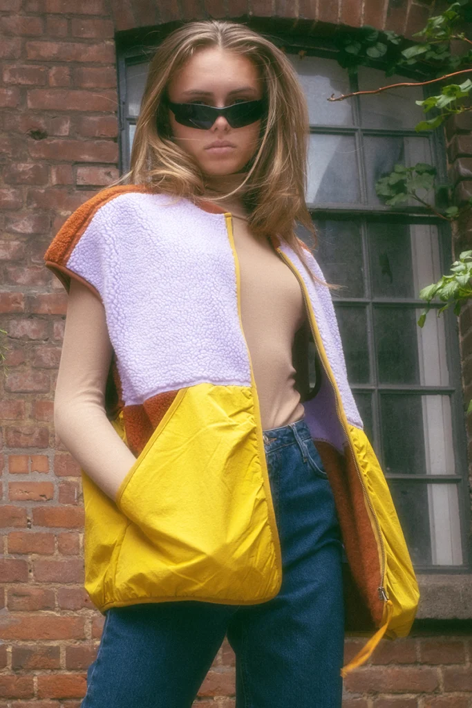 Model wearing a color-blocked fleece and nylon vest with sunglasses, standing in front of a brick wall.